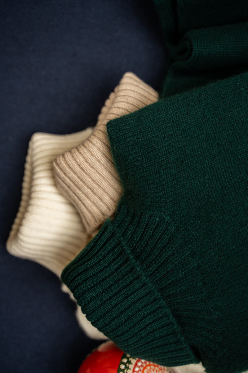 Close-up of folded knit garments in dark forest green and cream, showing ribbed cuffs and soft texture against a dark navy background.