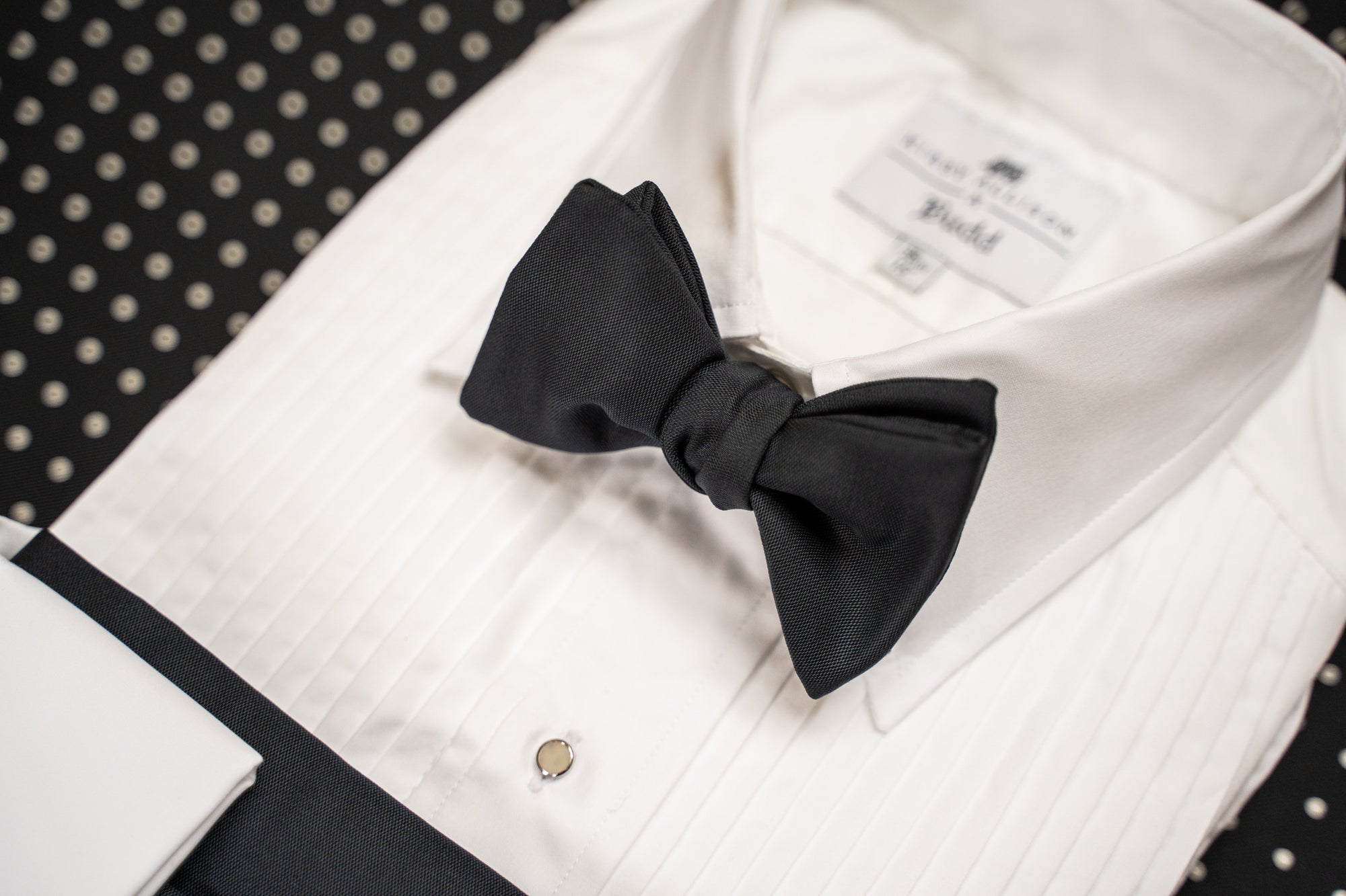 A black satin bow tie neatly tied on a white pinstriped dress shirt, with a black and white polka dot fabric background.