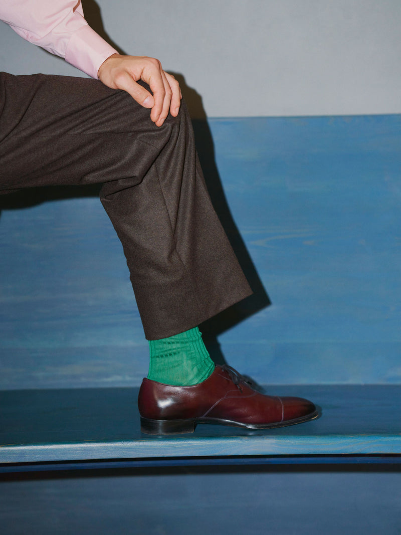 Man wearing trousers with his leg up on a blue bench showing his green socks and brown shoe, against a blue and white wall