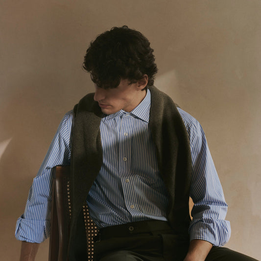 A man wearing a blue and white striped button-down shirt, a dark brown vest, and a patterned tie, seated against a warm beige wall.