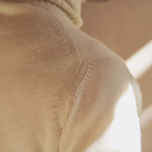 Close-up of a cream-colored knit turtleneck sweater with fine ribbed stitching, shown in warm natural light with a soft shadow.
