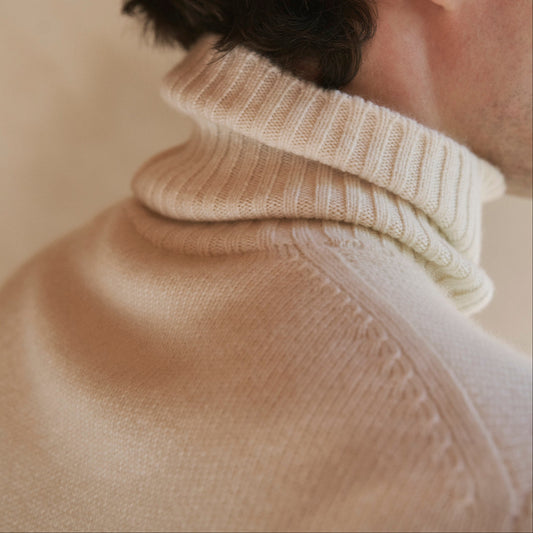 Close-up of a person wearing a cream ribbed turtleneck sweater against a warm beige background.