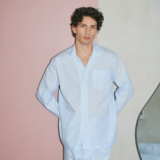 A man wearing a light blue long-sleeve button-up pyjama shirt and matching light blue trousers, standing against a split pink and off-white wall.