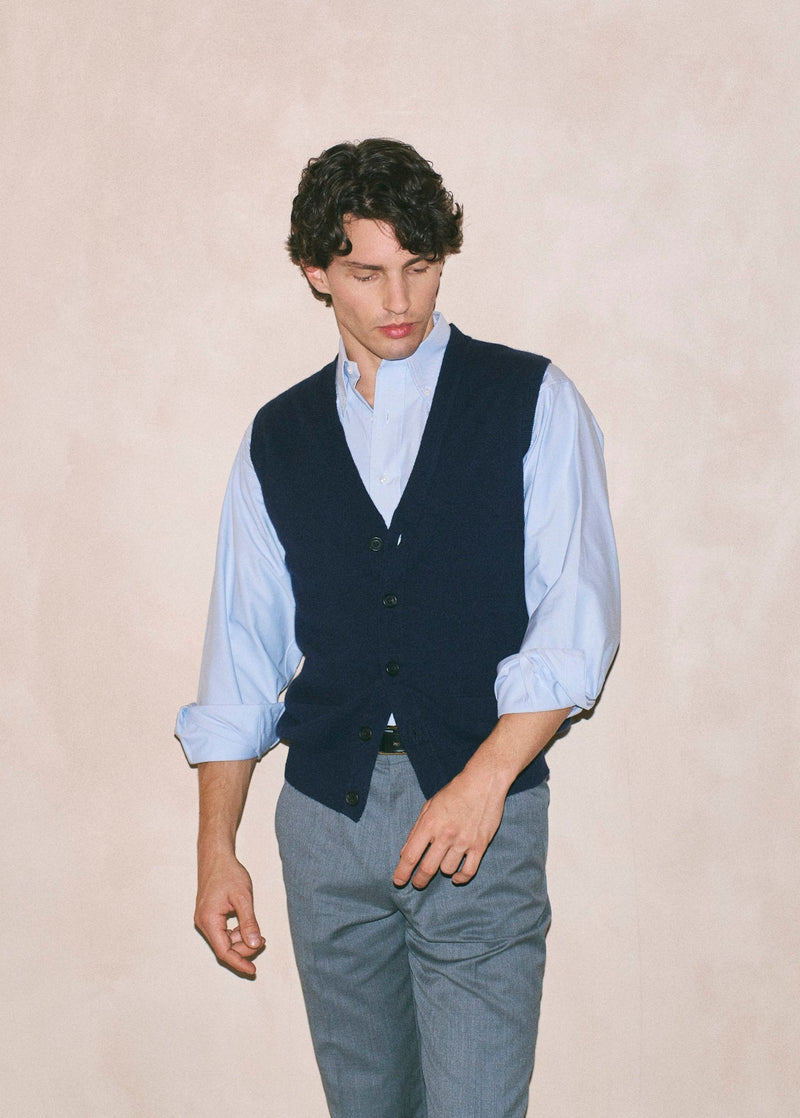 A man wearing a light blue long-sleeve button-up shirt, a dark navy vest, and gray trousers, standing against a textured beige wall.
