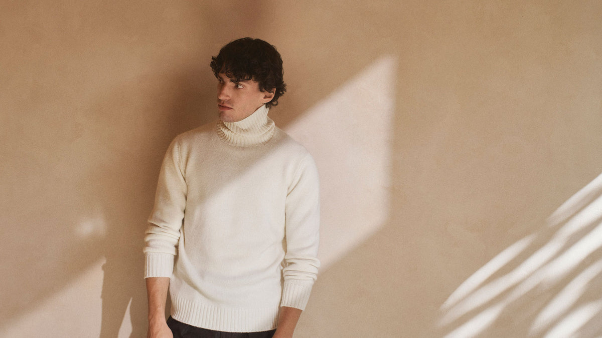 Model wearing cream cashmere roll neck knitted jumper stood against a neutral wall with dramatic sunlight shadows
