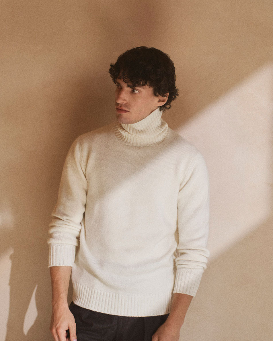 Model wearing a cashmere roll neck in cream stood against neutral wall with sunlight shadows across