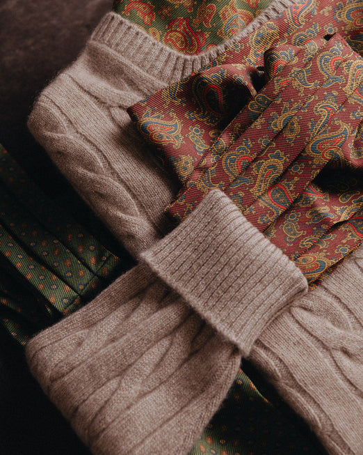 A close-up styled flat lay of coordinated fall fashion, featuring a chunky cable-knit sweater in taupe, a paisley necktie and matching patterned fabric in rust red, green, and gold, and dark green patterned material.