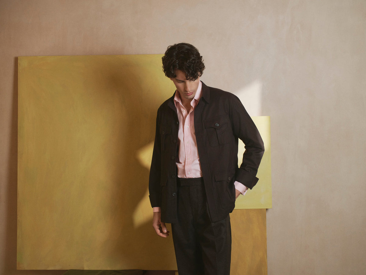 A man wearing a dark brown wool overshirt jacket over a light pink dress shirt and dark trousers, standing in front of a large yellow canvas painting against a beige wall.