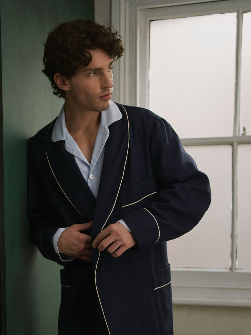 Man stood looking out of a window leant against a green wall wearing a navy dressing gown and blue pyjamas