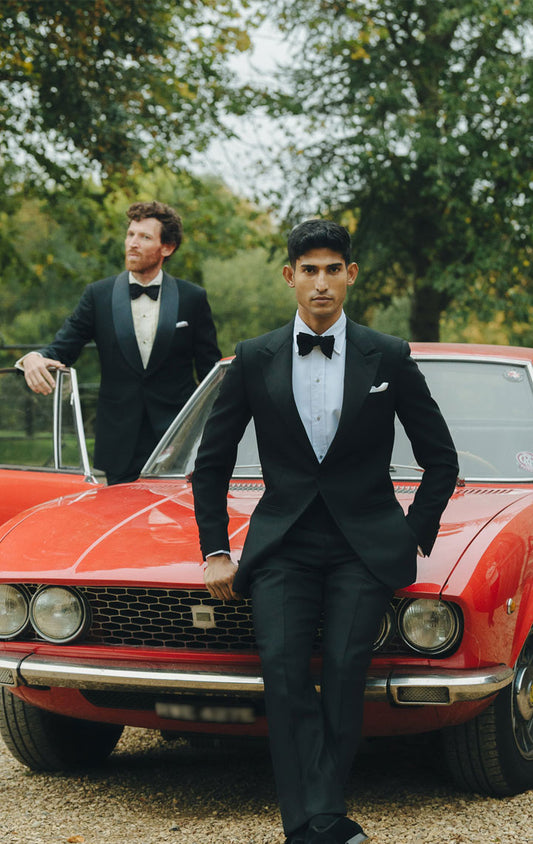 Two men in formal attire standing next to a red car with a stone building and greenery in the background.