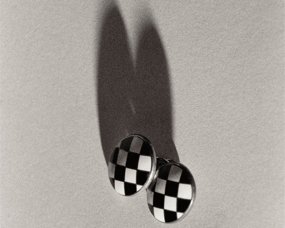 A pair of oval cufflinks with a black and white checkerboard pattern, photographed on a textured light gray background with a prominent shadow.