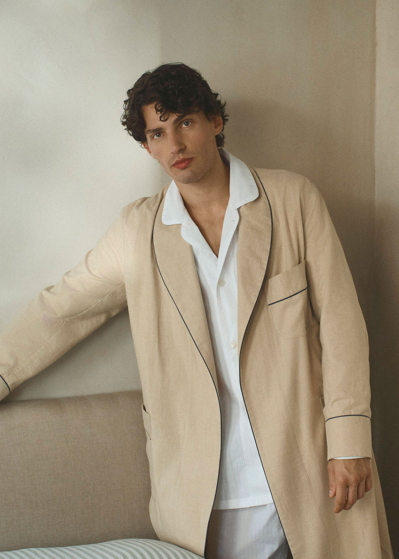 Man wearing a beige dressing gown over a white pyjama set stood by a neutral bedroom against a beige wall