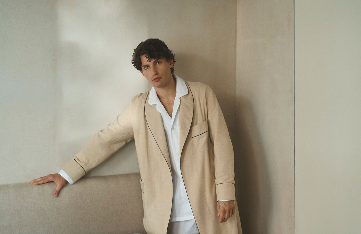 A man wearing a beige longline dressing gown over a white pyjama set, leaning against a neutral beige wall.