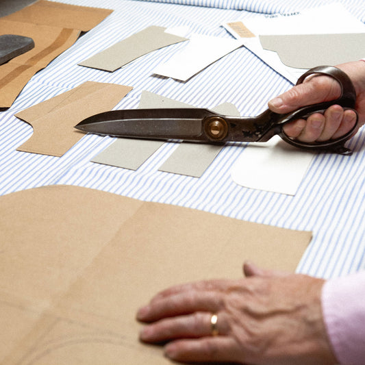 A close-up of a person's hands using large tailor's shears to cut brown cardboard or paper templates from a striped fabric tablecloth.