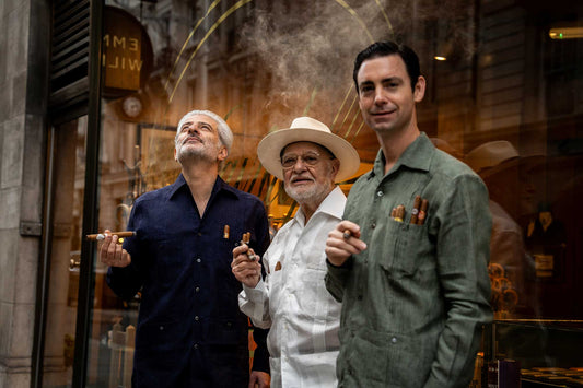 Three men standing together, with one wearing a hat, in front of a reflective surface.