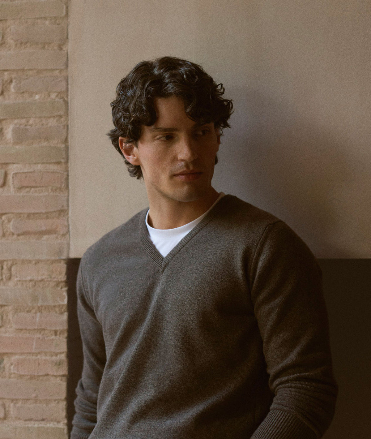 A man with dark curly hair wearing a dark gray v-neck sweater over a white t-shirt, leaning against a wall with a brick column on the left.