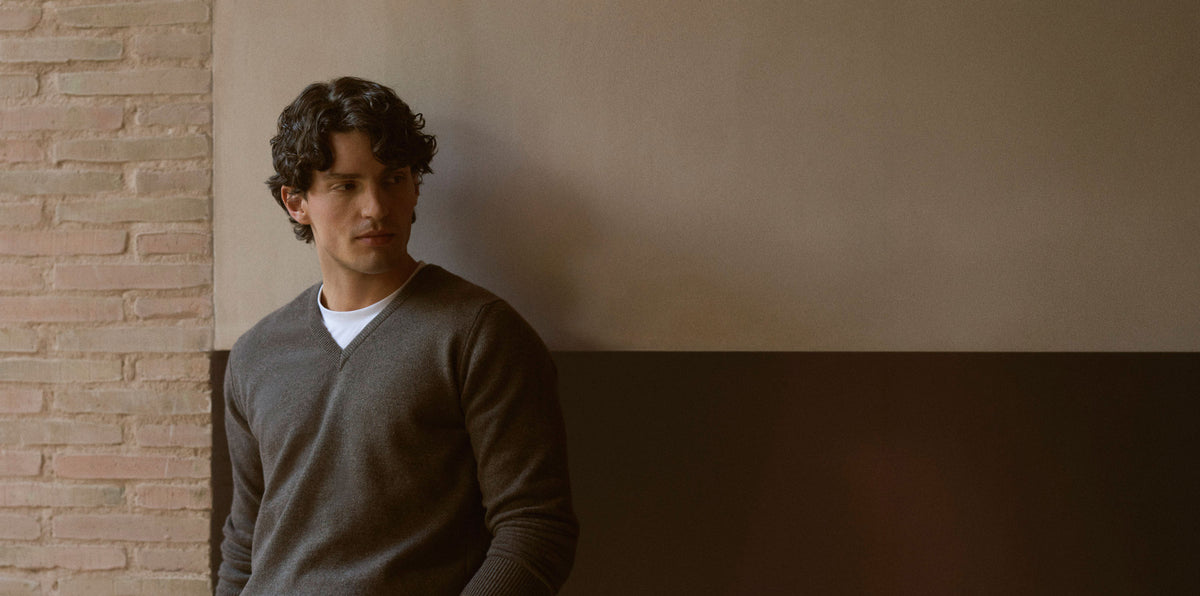 A man with curly dark hair wearing a dark gray v-neck sweater over a white undershirt, standing against a split brick and beige wall background.