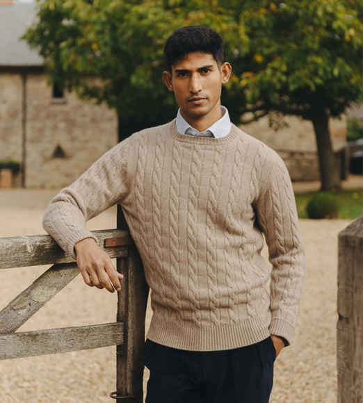 Man wearing a beige cable knit sweater standing outdoors near a wooden gate.