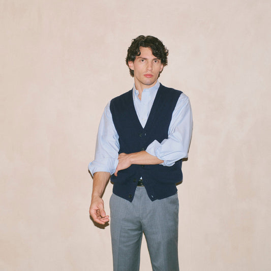 A man wearing a light blue dress shirt, a dark navy vest, and gray trousers, standing against a textured beige wall.