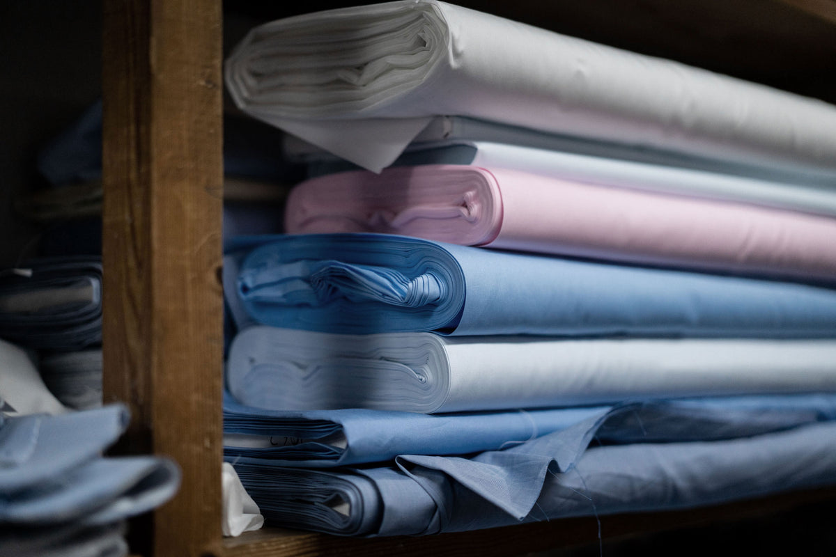 Stacks of folded and rolled fabric rolls in shades of blue, white, and pink, neatly arranged on a wooden shelf.