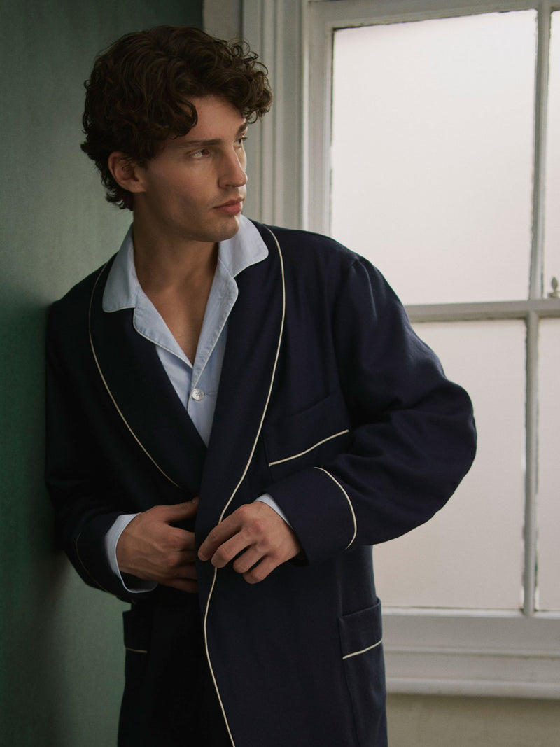 A man wearing a navy blue dressing gown with white piping over a light blue button-down shirt, leaning against a dark green wall next to a window.