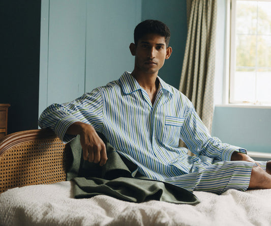 Man in a striped night shirt sitting on a bed in a room with a window and curtains.