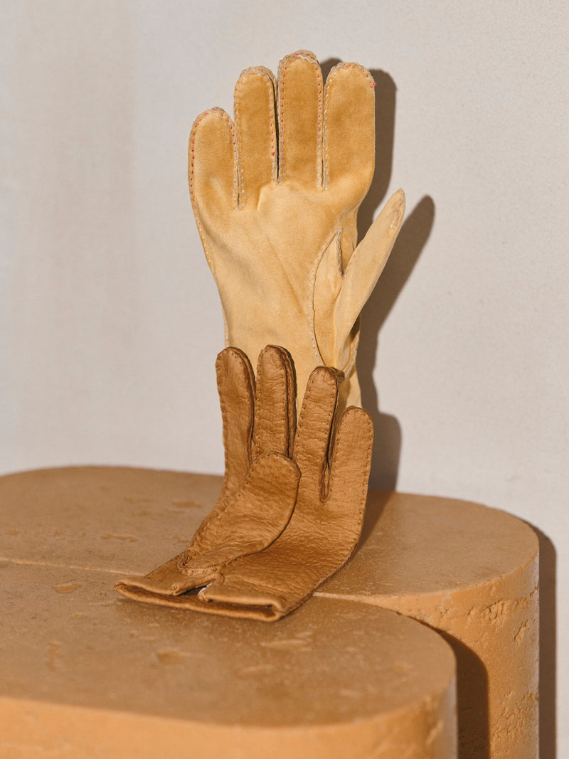 Two tan leather gloves, one larger and one smaller, arranged upright on a textured tan pedestal against a light beige wall.