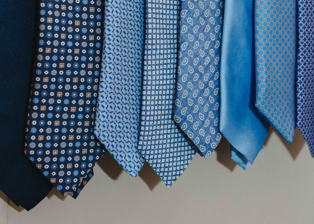 A close-up row of blue neckties hanging side by side, featuring different patterns including polka dots, small geometric shapes, and solid fabric in shades of navy, medium blue, and light blue.