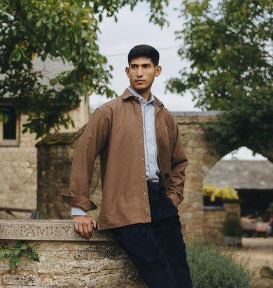 Man in a brown jacket standing outdoors with stone building and trees in the background
