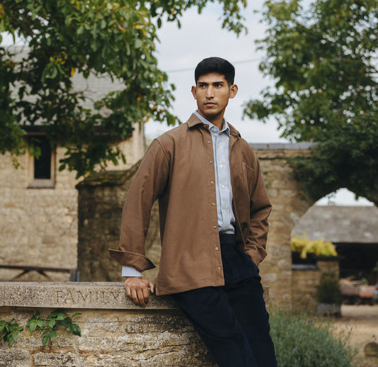 Man in a brown jacket standing outdoors with stone building and trees in the background
