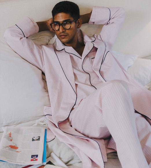 Man wearing pink pajamas with black piping, sitting on a bed.