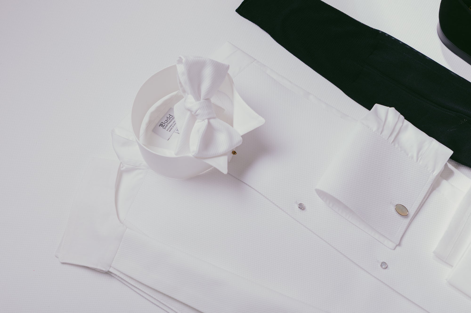 A styled flat-lay of a white long-sleeve shirt with a large white bow on the collar, a white cuff with a gold button, and a dark green fabric accessory on a white background.