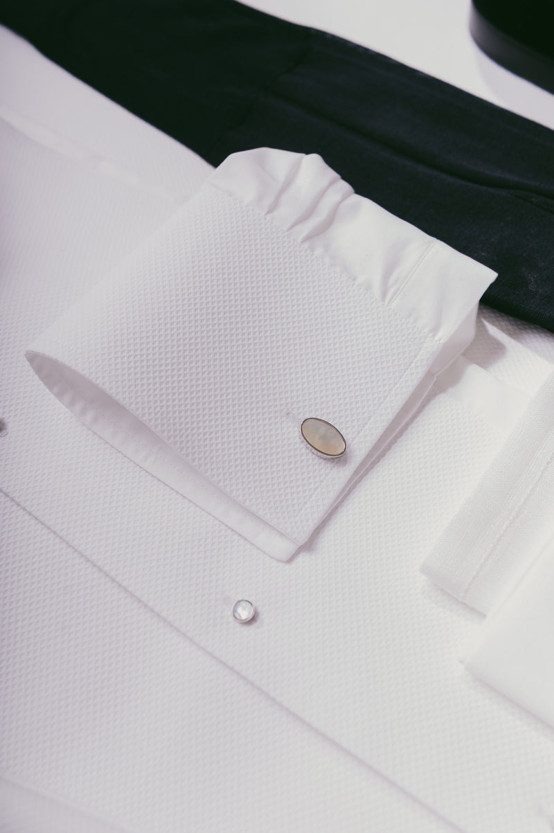 Close-up of a white textured dress shirt cuff with a silver oval cufflink, laid over a white shirt body with a black tie partially visible behind it.