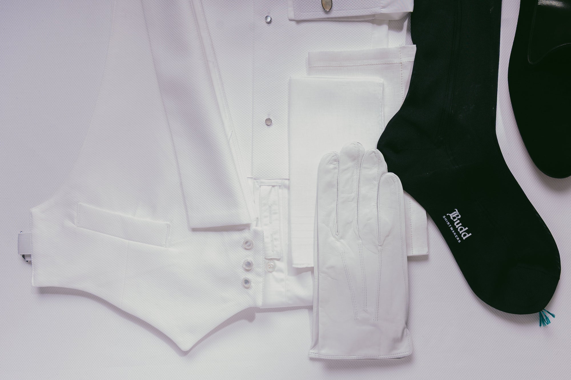 A styled flat-lay of formal wear items on a white background, including a white vest, white shirt, white gloves, black socks, and black dress shoes. The socks have a visible white 'Bucci' logo.