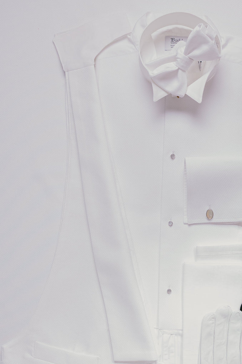 A flat-lay of a white formal outfit including a long white shirt, white vest, white bow tie, white pocket square, and a single black sock, arranged on a white background.