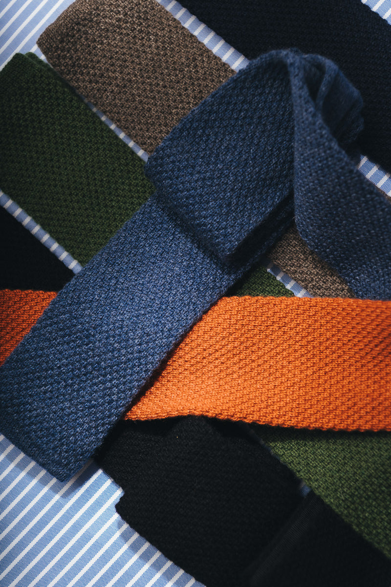 A close-up of several knitted neckties arranged on a striped fabric background. The most prominent tie is dark blue, with other visible ties in olive green, brown, orange, black, and navy.