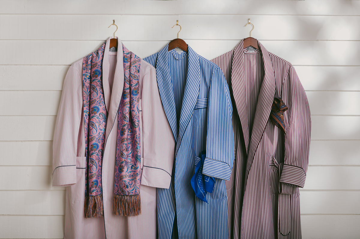 Three elegant robes hanging on wooden hangers against a white shiplap wall, styled with a paisley scarf. The left robe is light pink with a multicolor paisley scarf.