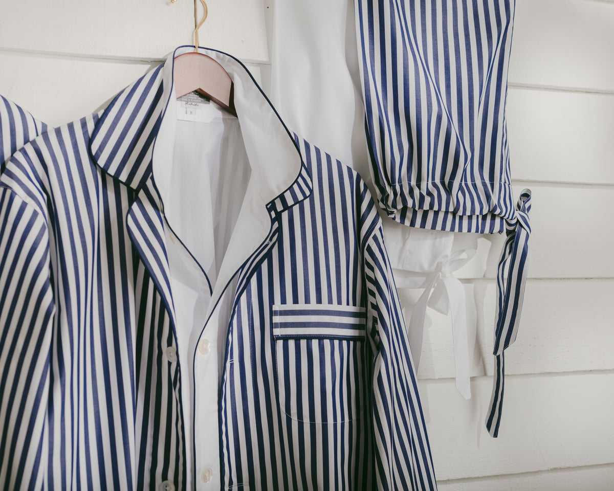 A matching pajama set with blue and white vertical stripes, featuring a long-sleeve button-front top with a collar and a pair of coordinating tie-waist pants, hanging against a white paneled wall.