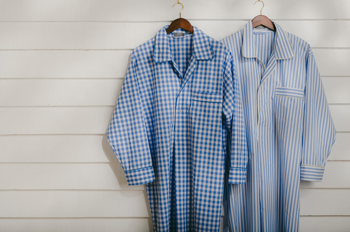 Two long blue-and-white check and striped pajama shirts hanging on wooden hangers against a white wood plank wall.