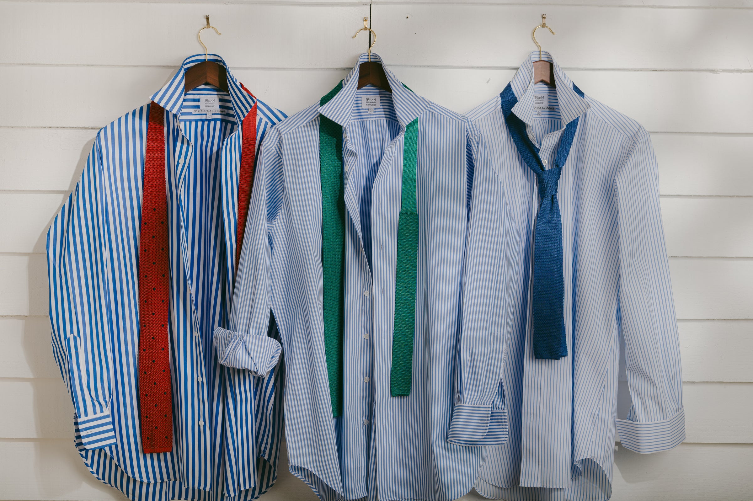 Three striped shirts with suspenders and ties on hangers against a white wooden panel background