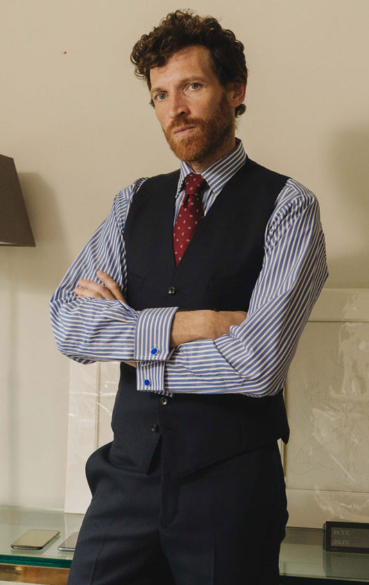 Man in a striped shirt and vest standing in a room with framed pictures and a lamp.