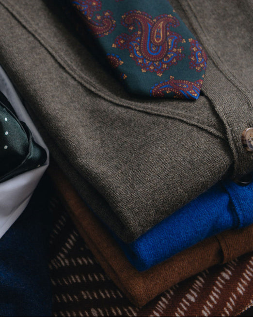 A folded stack of men's knit clothing and ties, featuring a dark teal paisley necktie with red and gold detailing, a brown wool sweater, a bright blue knit layer, and a brown patterned fabric underneath.