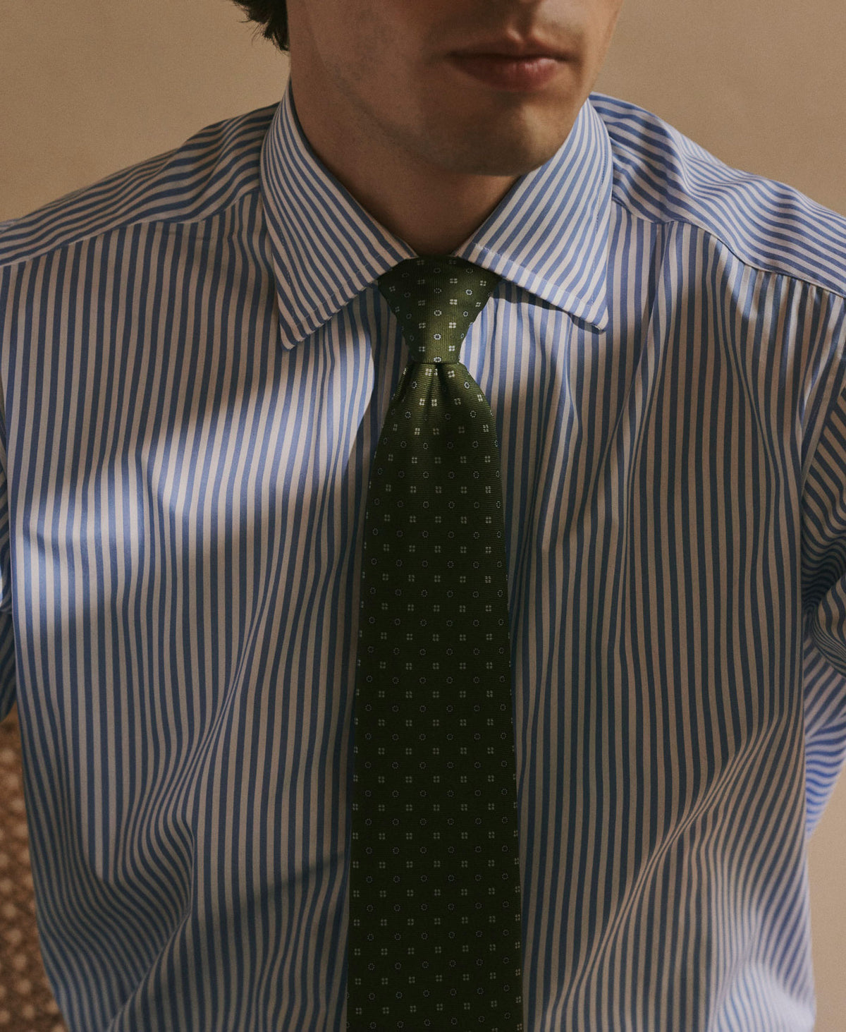 A man wearing a blue and white vertically striped dress shirt with a dark green patterned tie, shown from the chest up against a warm beige background.