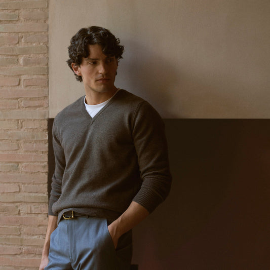 A man wearing a dark gray v-neck sweater over a white undershirt and blue trousers, leaning against a wall in a studio setting with a brick wall on the left.